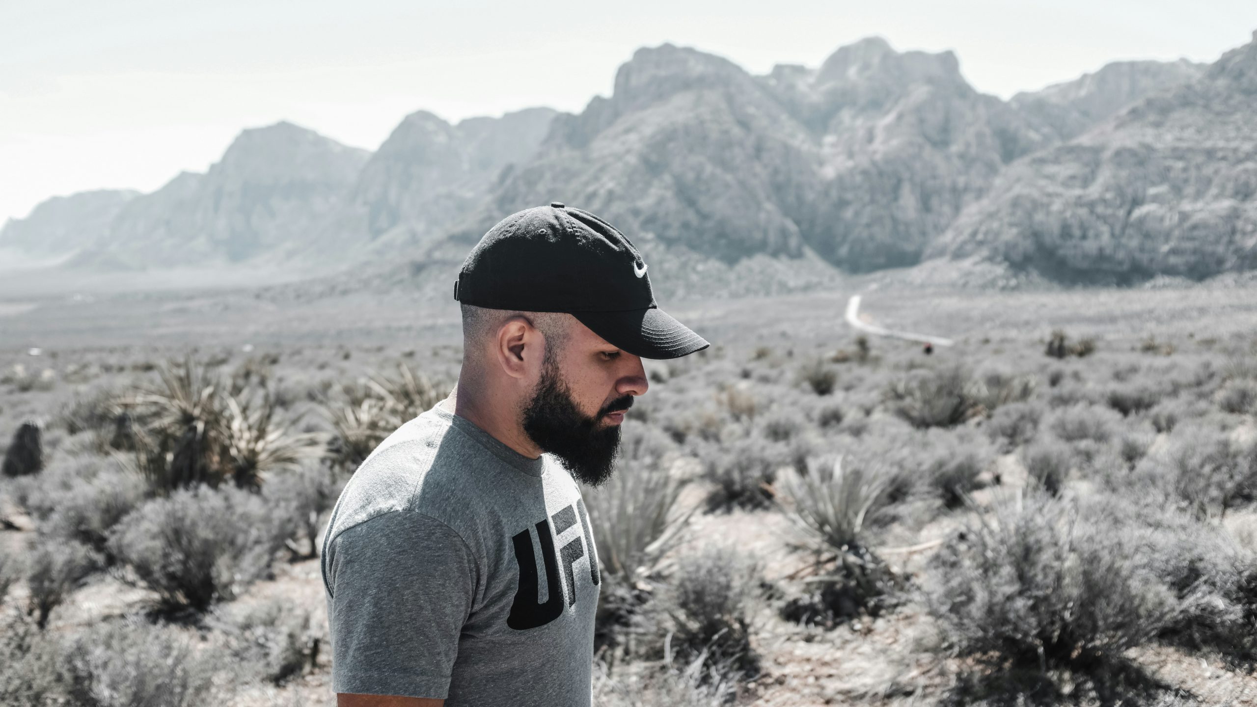 Man with a black cap and gray T‑shirt standing in a dry desert landscape with rocky mountains in the background (profile view).
