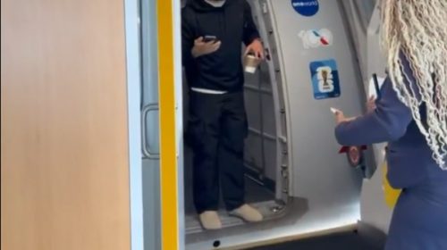 Man in a black hoodie stands in an airplane doorway, holding a cup in one hand and a phone in the other, while a woman with long curly hair approaches from outside the cabin.