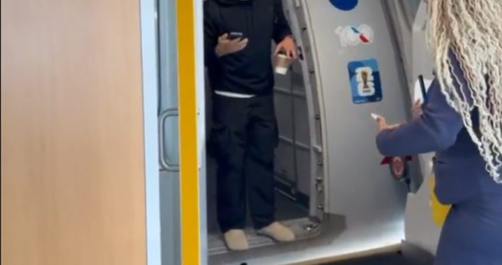 Man in a black hoodie stands in an airplane doorway, holding a cup in one hand and a phone in the other, while a woman with long curly hair approaches from outside the cabin.