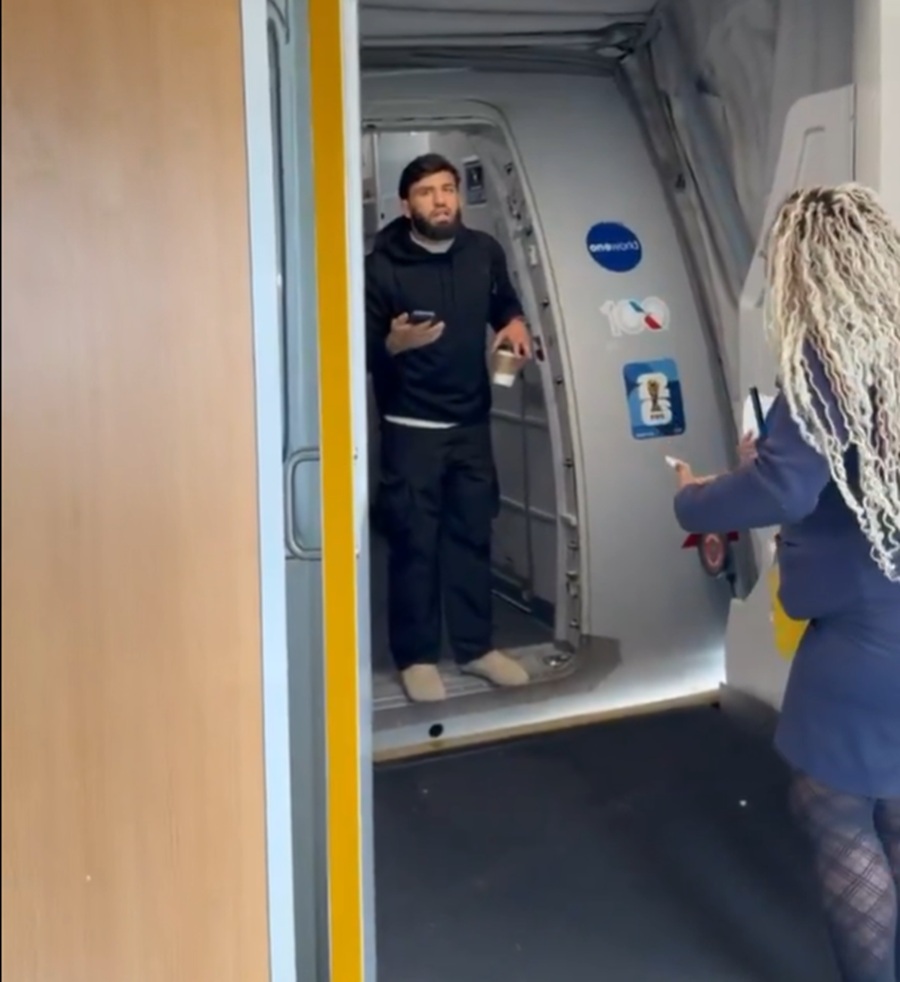 Man in a black hoodie stands in an airplane doorway, holding a cup in one hand and a phone in the other, while a woman with long curly hair approaches from outside the cabin.