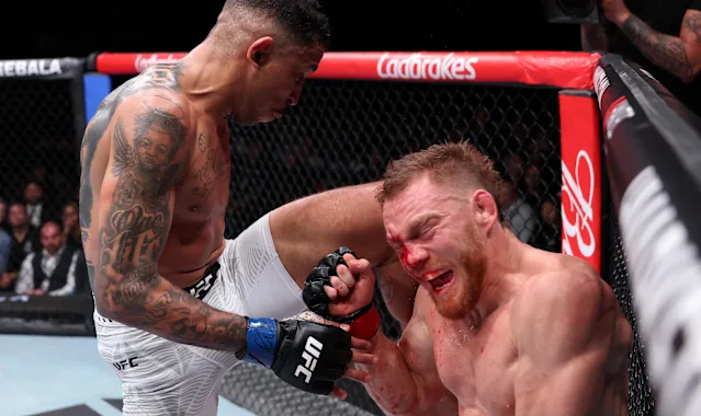 Two UFC fighters in the octagon: one lands a knee to the other's face, both battered and bleeding, with a camera crew and spectators in the background.