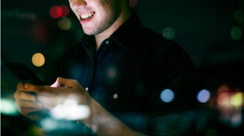Man in a dark shirt smiles while looking at a smartphone at night, city lights blurred in the background.