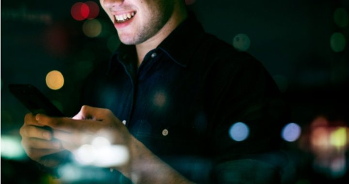 Man in a dark shirt smiles while looking at a smartphone at night, city lights blurred in the background.