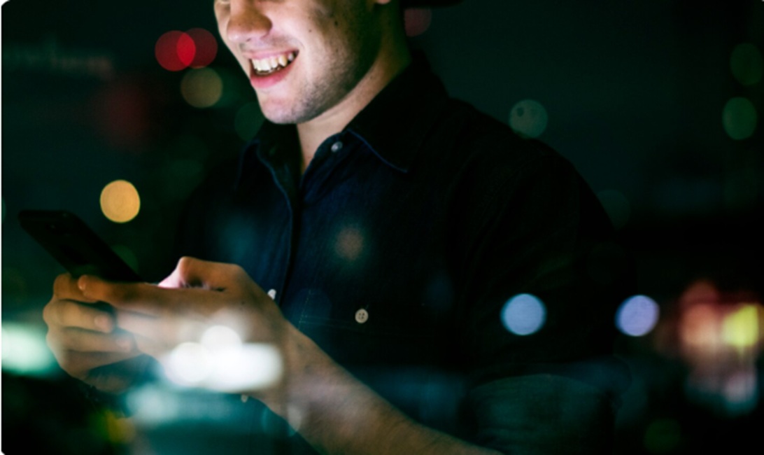 Man in a dark shirt smiles while looking at a smartphone at night, city lights blurred in the background.
