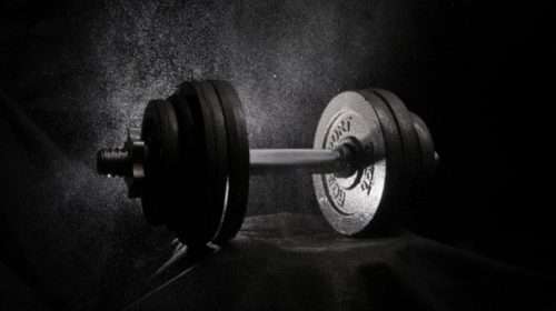 Dumbbell with several weight plates on a bar resting on a dark surface, lit from the side with dust particles in the air.