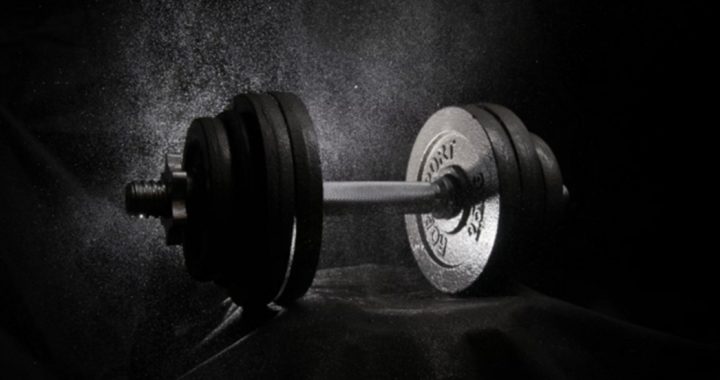 Dumbbell with several weight plates on a bar resting on a dark surface, lit from the side with dust particles in the air.