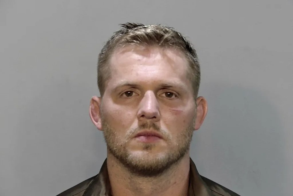 Mugshot of a man with short blond hair and stubble facing the camera against a gray background.