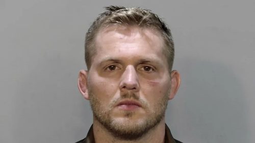 Mugshot of a man with short blond hair and stubble facing the camera against a gray background.