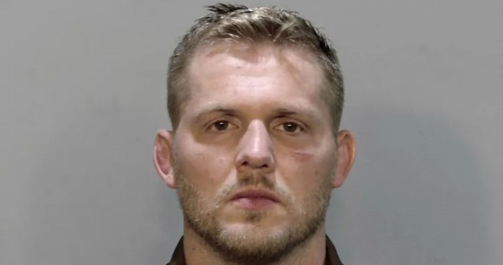 Mugshot of a man with short blond hair and stubble facing the camera against a gray background.