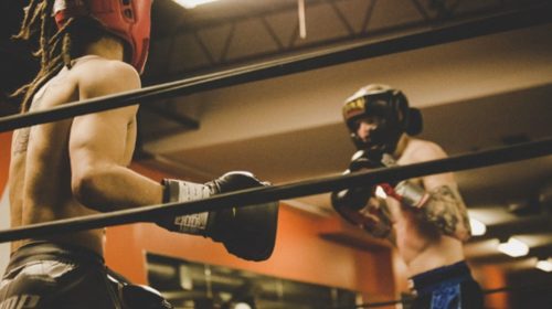 Two boxers spar in a gym ring; one in red headgear, the other in blue shorts and black gloves._Turns into a concise description of action and context.