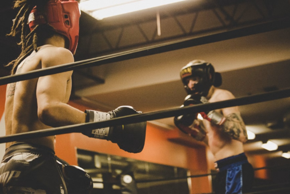 Two boxers spar in a gym ring; one in red headgear, the other in blue shorts and black gloves._Turns into a concise description of action and context.