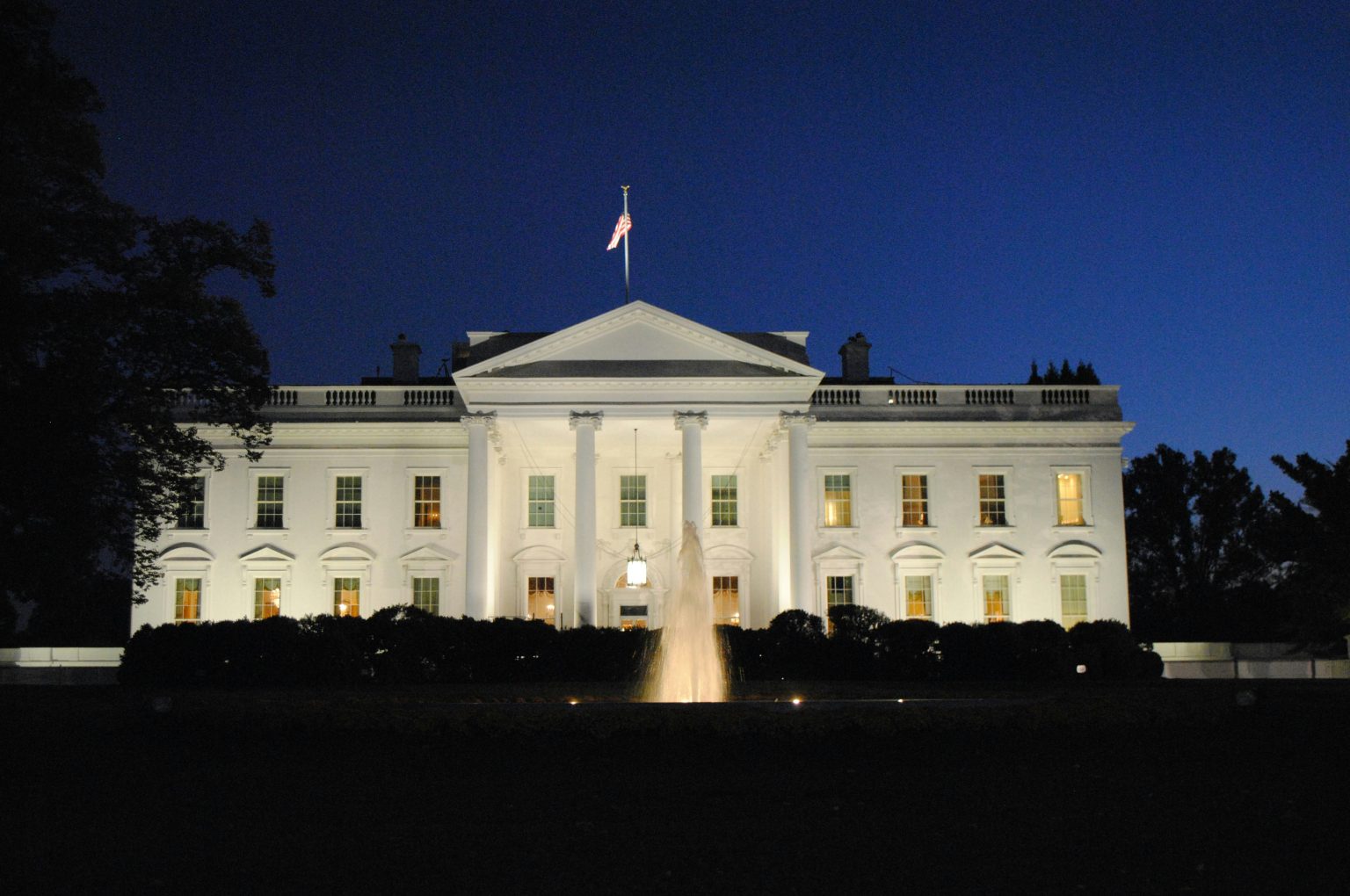 White neoclassical government building at night with an American flag atop and a lit fountain in front.