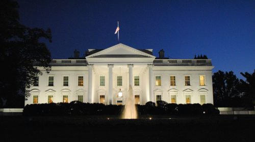 White neoclassical government building at night with an American flag atop and a lit fountain in front.