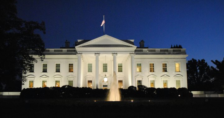 White neoclassical government building at night with an American flag atop and a lit fountain in front.