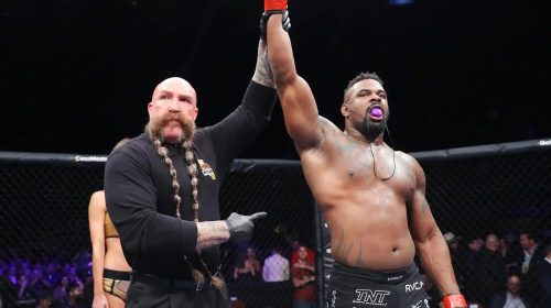 MMA referee raises the winner's arm as a shirtless fighter with a purple mouthguard celebrates inside the cage.