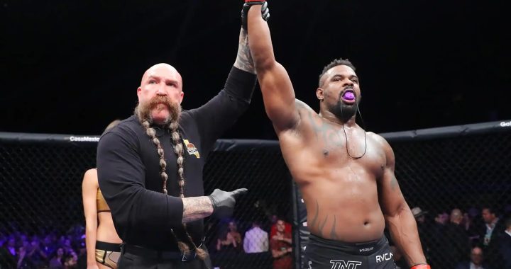 MMA referee raises the winner's arm as a shirtless fighter with a purple mouthguard celebrates inside the cage.