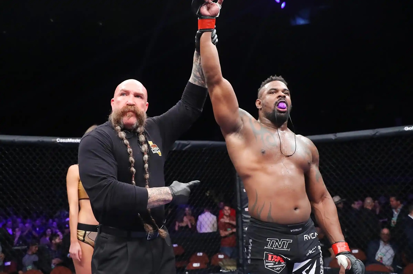 MMA referee raises the winner's arm as a shirtless fighter with a purple mouthguard celebrates inside the cage.