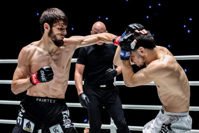 Two shirtless fighters exchange punches in a ring while a referee watches in the background.
