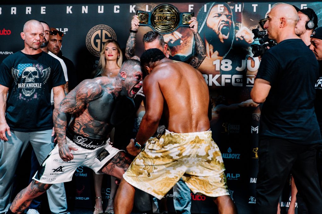 Two muscular fighters face off in a staredown, one shirtless with tatts, the other in camo shorts, during a weigh-in event with a belt held overhead behind them