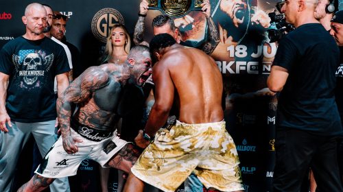 Two muscular fighters face off in a staredown, one shirtless with tatts, the other in camo shorts, during a weigh-in event with a belt held overhead behind them