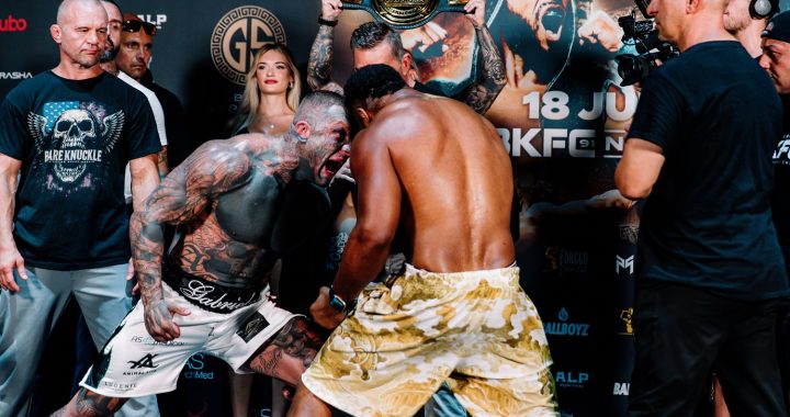 Two muscular fighters face off in a staredown, one shirtless with tatts, the other in camo shorts, during a weigh-in event with a belt held overhead behind them