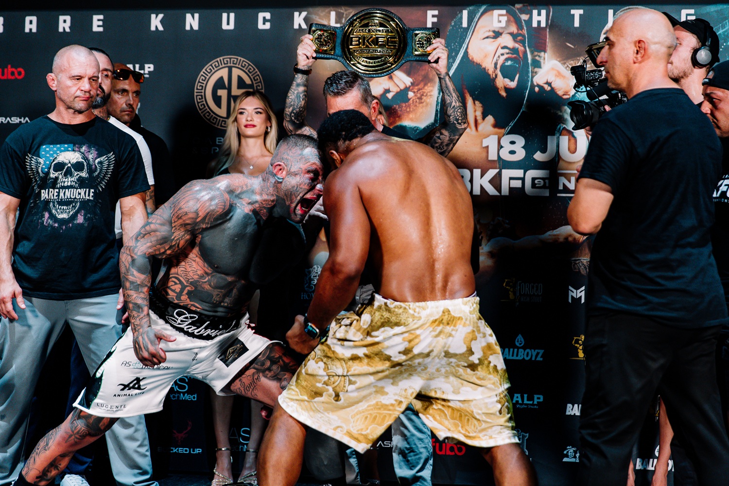 Two muscular fighters face off in a staredown, one shirtless with tatts, the other in camo shorts, during a weigh-in event with a belt held overhead behind them