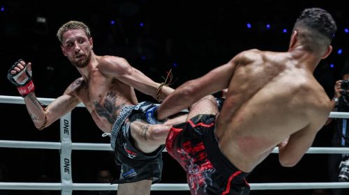 Two shirtless fighters clash in a ring; one lands a high kick to the other while wearing red gloves and black shorts with a floral pattern.