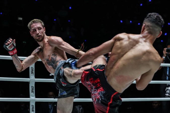 Two shirtless fighters clash in a ring; one lands a high kick to the other while wearing red gloves and black shorts with a floral pattern.