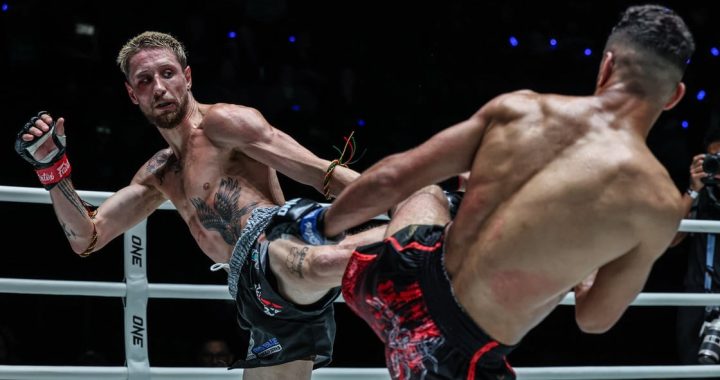 Two shirtless fighters clash in a ring; one lands a high kick to the other while wearing red gloves and black shorts with a floral pattern.