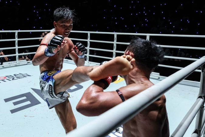 Two male mixed martial arts fighters in a ring; the left fighter delivers a kick as the opponent guards with raised arms.