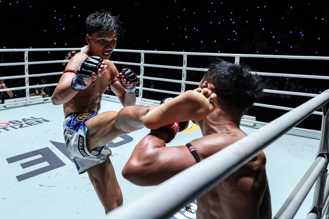 Two male mixed martial arts fighters in a ring; the left fighter delivers a kick as the opponent guards with raised arms.