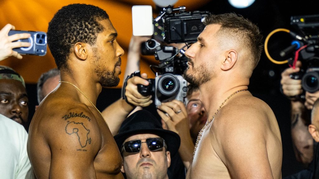 Two shirtless men face off in a stare-down as photographers circle around them during a weigh‑in or press event, one with a tattoo of Africa and the word 'Wisdom' on his shoulder