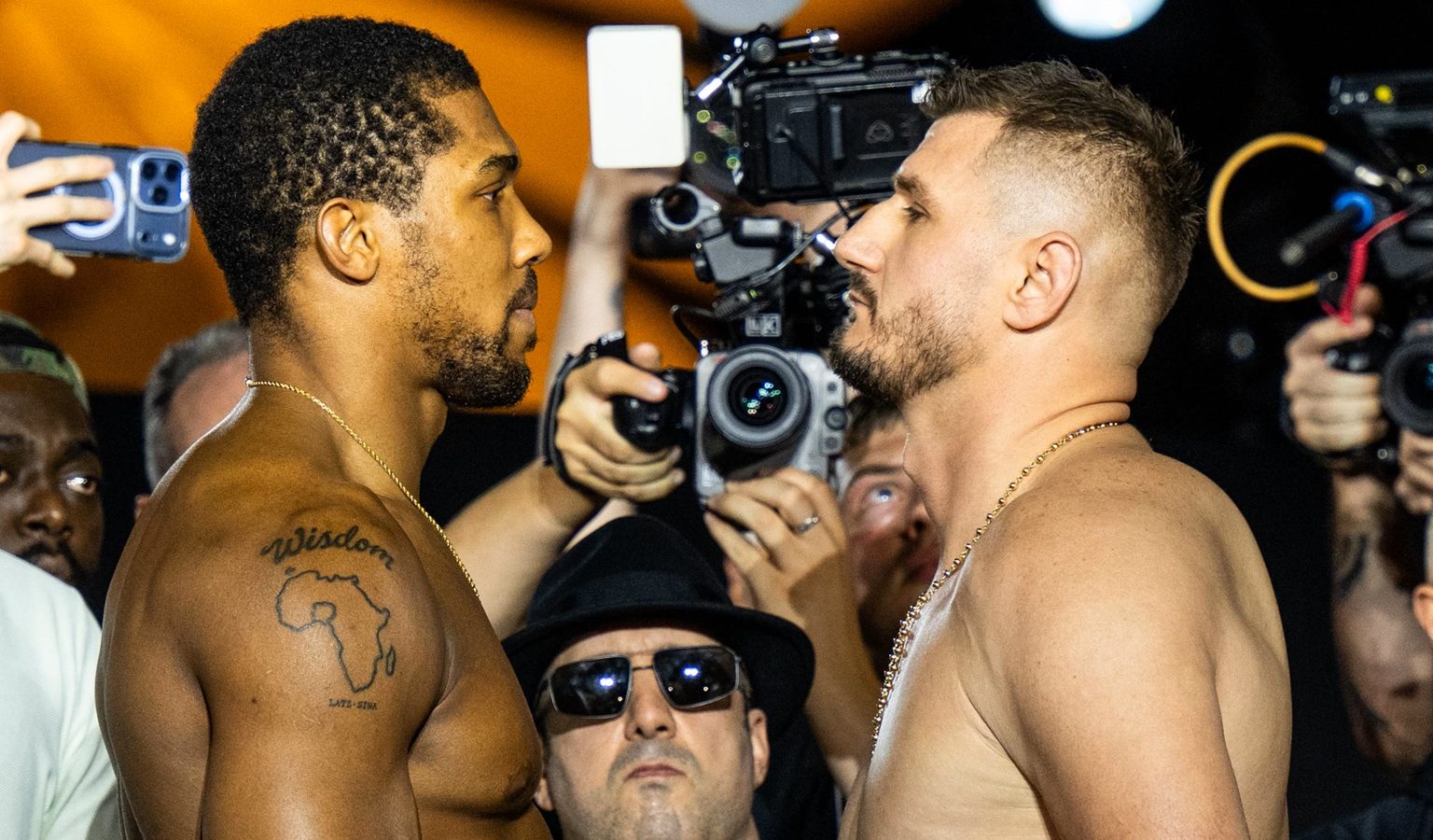 Two shirtless men face off in a stare-down as photographers circle around them during a weigh‑in or press event, one with a tattoo of Africa and the word 'Wisdom' on his shoulder