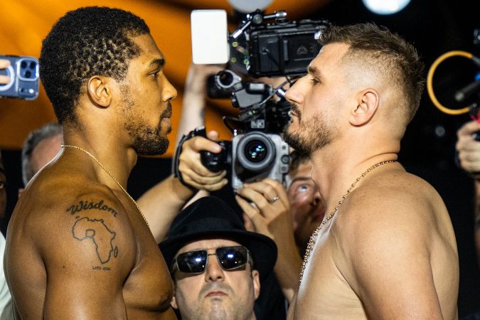Two shirtless men face off in a stare-down as photographers circle around them during a weigh‑in or press event, one with a tattoo of Africa and the word 'Wisdom' on his shoulder