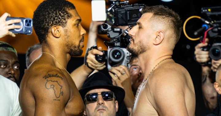 Two shirtless men face off in a stare-down as photographers circle around them during a weigh‑in or press event, one with a tattoo of Africa and the word 'Wisdom' on his shoulder