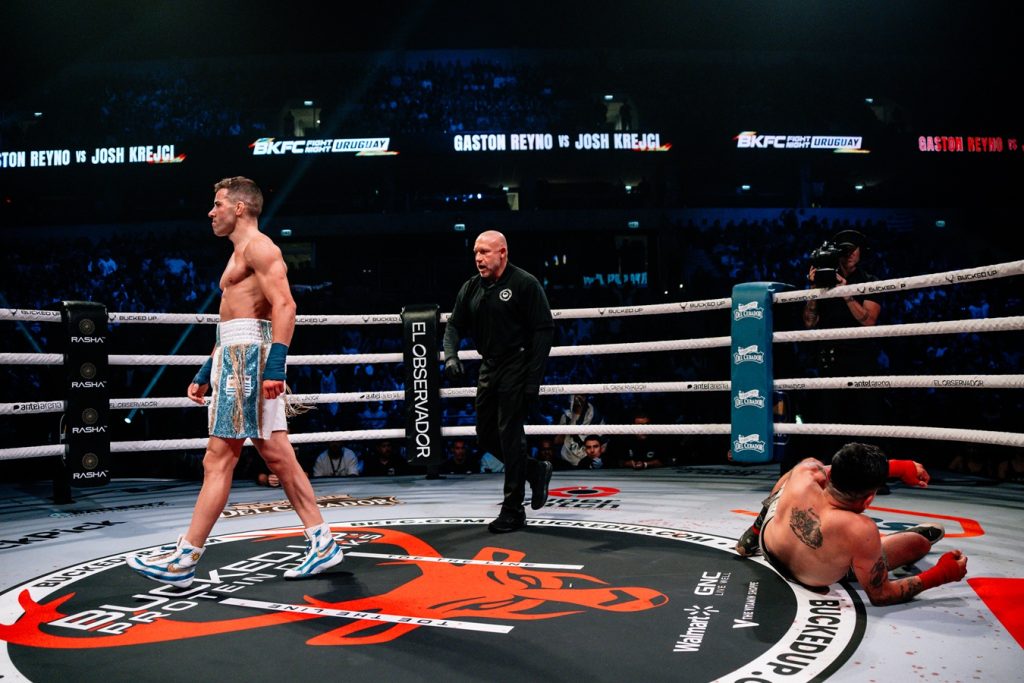 Professional boxing ring scene: one fighter standing in blue shorts and gloves, another fighter on the mat, with the referee approaching and a cheering crowd in the background.