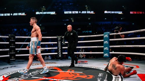 Professional boxing ring scene: one fighter standing in blue shorts and gloves, another fighter on the mat, with the referee approaching and a cheering crowd in the background.