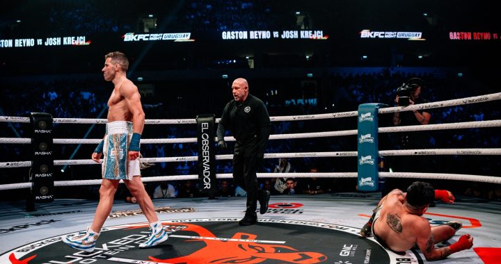 Professional boxing ring scene: one fighter standing in blue shorts and gloves, another fighter on the mat, with the referee approaching and a cheering crowd in the background.