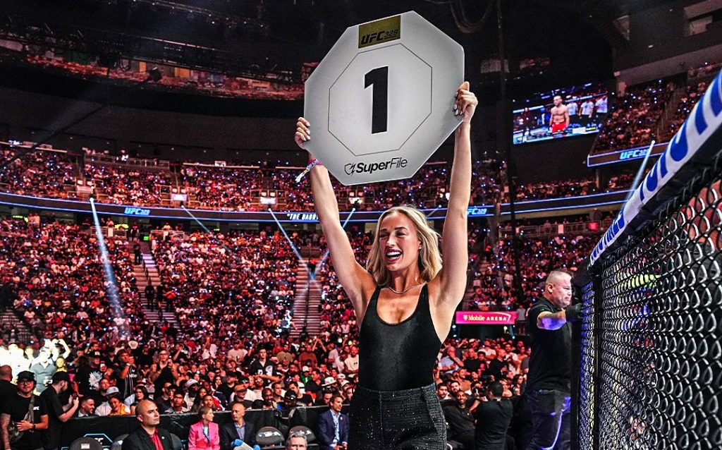 Blonde woman in a black outfit raises a large placard that shows the number 1 in a packed UFC arena, celebrating a win.