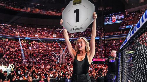 Blonde woman in a black outfit raises a large placard that shows the number 1 in a packed UFC arena, celebrating a win.