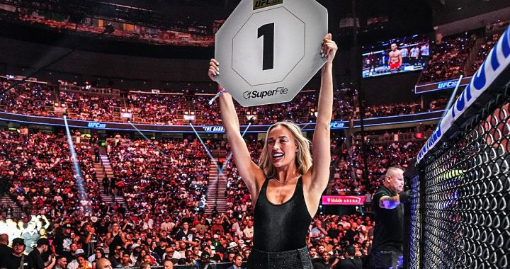 Blonde woman in a black outfit raises a large placard that shows the number 1 in a packed UFC arena, celebrating a win.